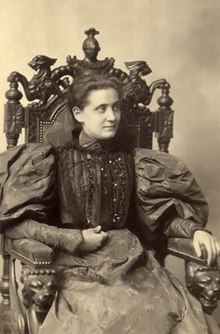 A woman in a Victorian-era dress with puffed sleeves sits on an ornate chair with carved griffins, looking slightly to the side. The photo is black and white, with a formal, historical atmosphere.
