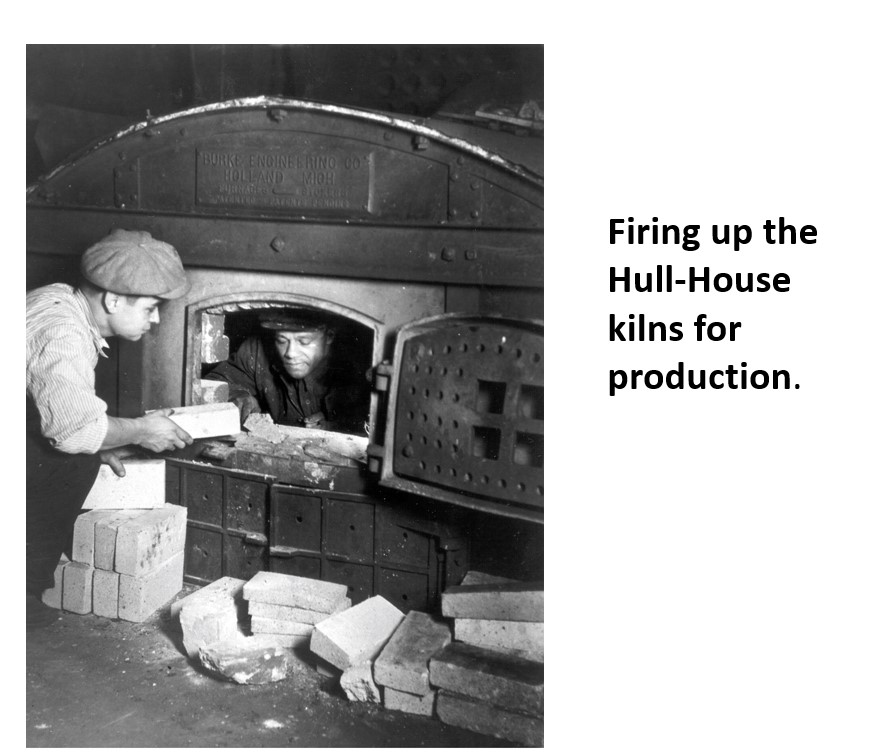 Two people place bricks into a large kiln at Hull-House, preparing it for production. Stacks of bricks are in front of the kiln, and one person is looking inside while the other hands over a brick.