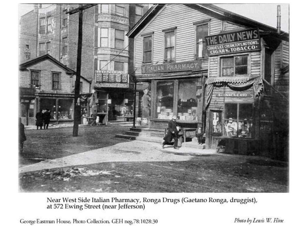 Black-and-white photo of people outside an Italian pharmacy and drugstore on a street corner, with signs for cigars and newspapers. Early 1900s urban setting with surrounding buildings and a man sitting on a bench.