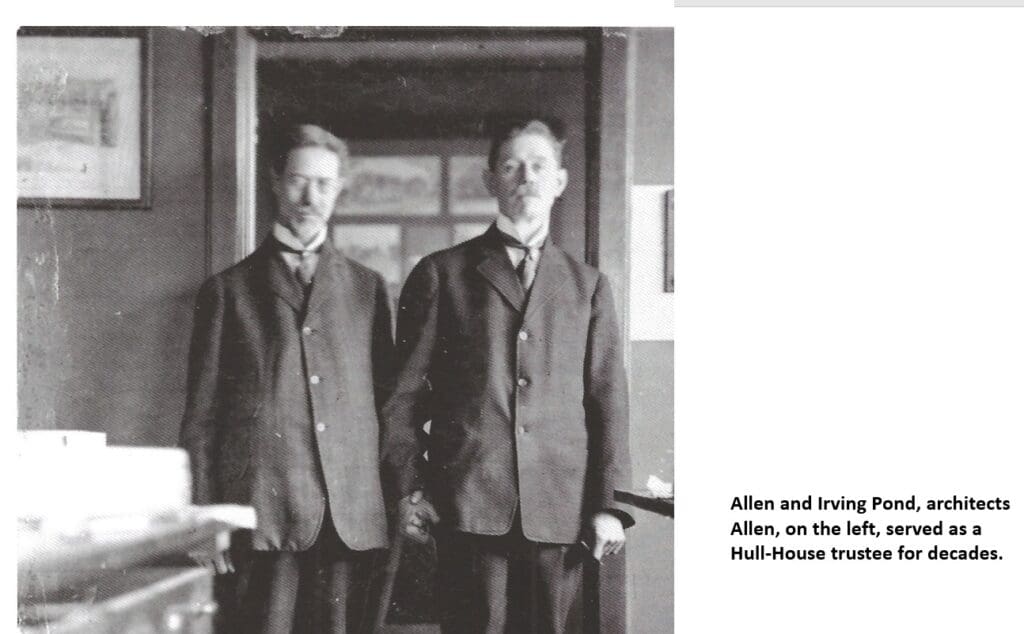 Two men in suits stand side by side in an old black-and-white photo. They are indoors with framed pictures on the wall behind them. Both look at the camera, with one slightly blurred.