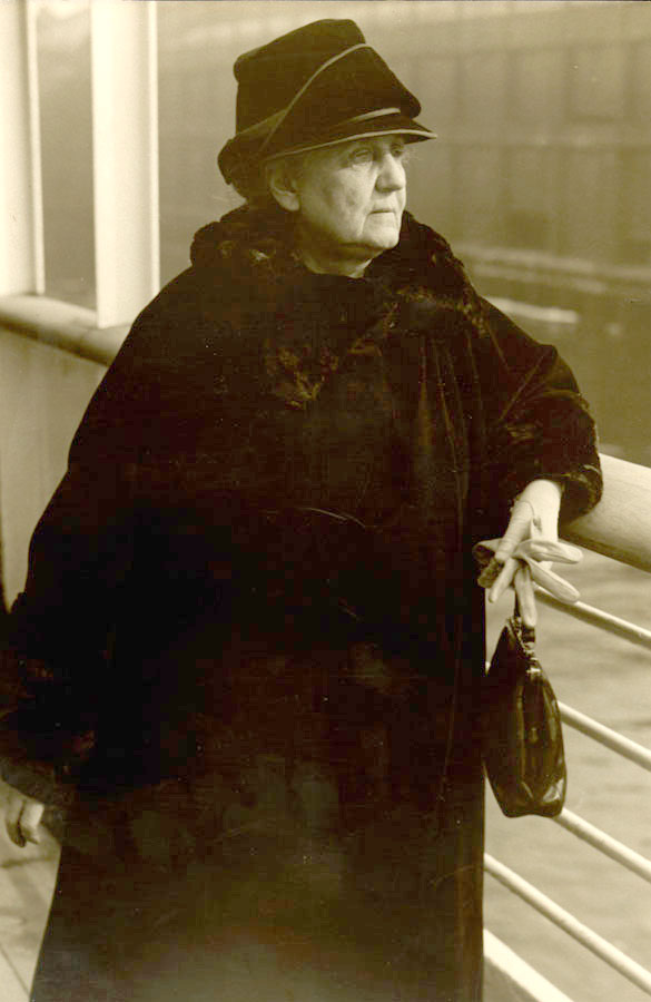 Jane Addams in a dark fur coat and hat stands by a railing, holding a small handbag and gloves, looking thoughtfully into the distance. The image is in sepia tone.