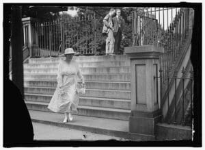 A woman in a long white dress and wide-brimmed hat descends outdoor stone steps, holding her skirt. Two men in suits stand at the top of the steps, watching her. Wrought iron railings line the stairs.