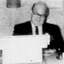 A black-and-white, grainy photo of a man in a suit and glasses sitting behind a desk or podium, looking at the camera.