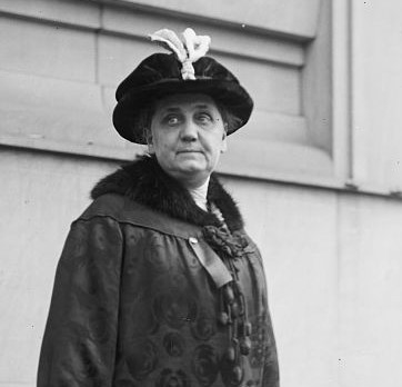 A woman wearing a dark, patterned coat with a fur collar and a decorative hat stands in front of a building, looking slightly to the side.
