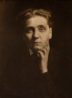 A sepia-toned portrait of a woman with short hair, gazing thoughtfully at the camera, her hand resting under her chin. She wears dark clothing and appears pensive against a plain background.