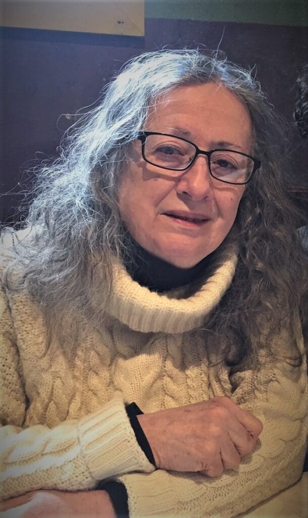 An older woman with long gray hair and glasses, wearing a cream-colored cable-knit turtleneck sweater, sits indoors at a table, softly smiling at the camera.