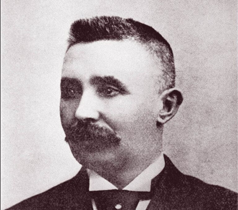 A sepia-toned portrait of a man with short hair and a thick mustache, wearing a suit and bow tie, looking slightly to his left.