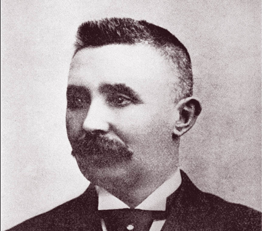 A sepia-toned portrait of a man with short hair and a thick mustache, wearing a suit and bow tie, looking slightly to his left.