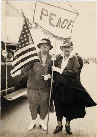 Two women in dresses and hats stand beside a car, holding an American flag and a banner reading PEACE. The photo is black and white, and a few people are visible in the background.